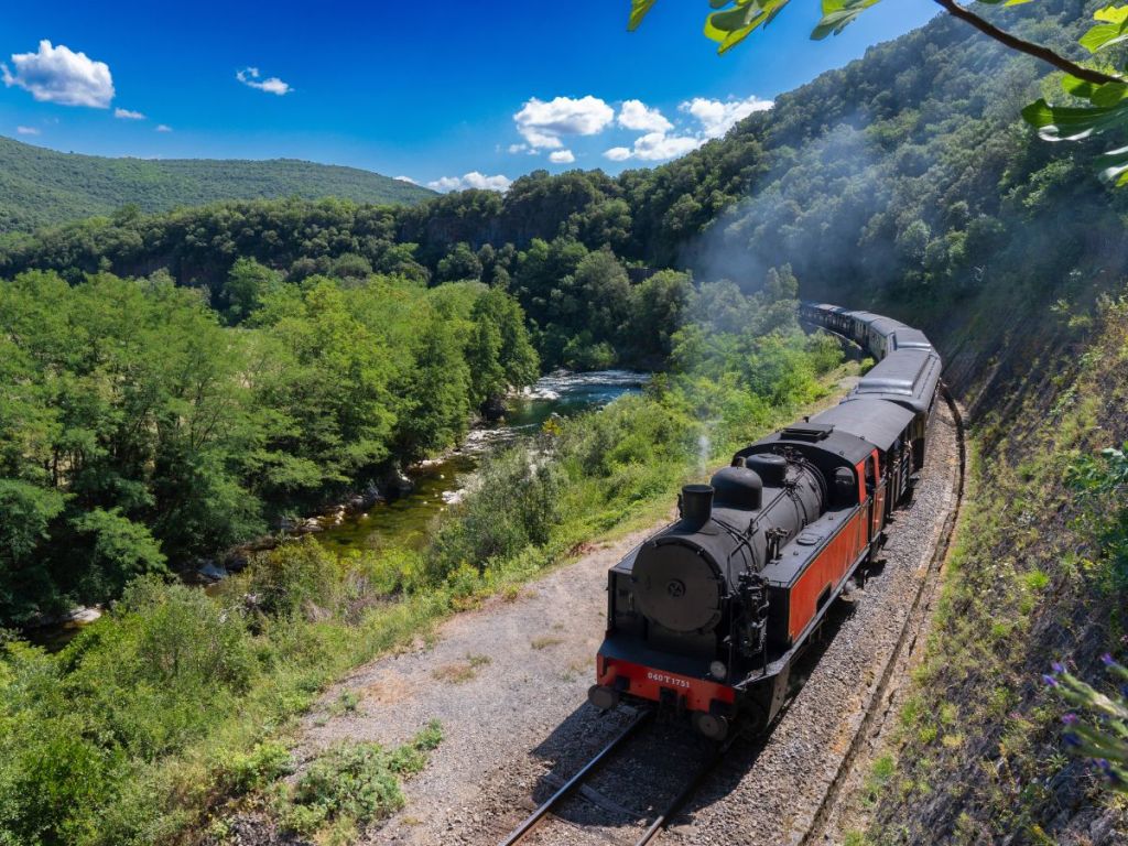Vintage steam locomotive of the Train des Cévennes winding through a lush river gorge in the Gard department of southern France