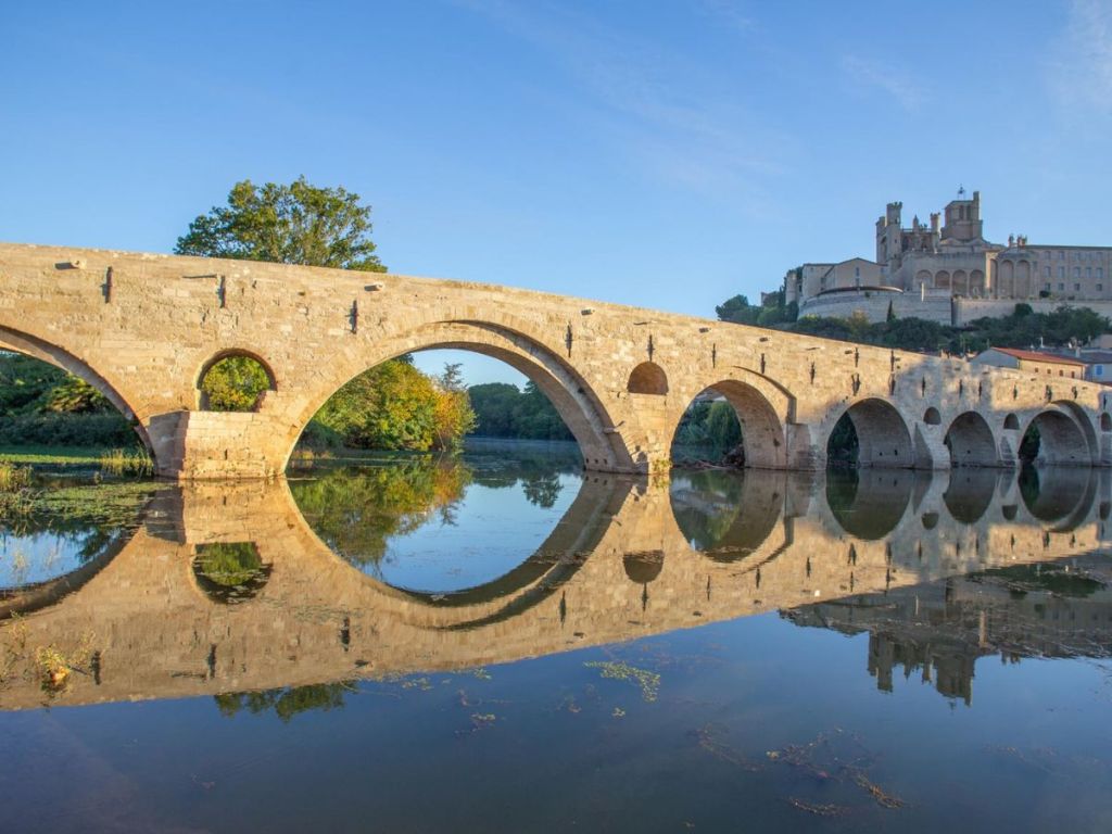 The medieval Pont Vieux of Béziers with its rounded stone arches perfectly reflected in the still waters of the Orb river, with Saint-Nazaire Cathedral crowning the hill behind