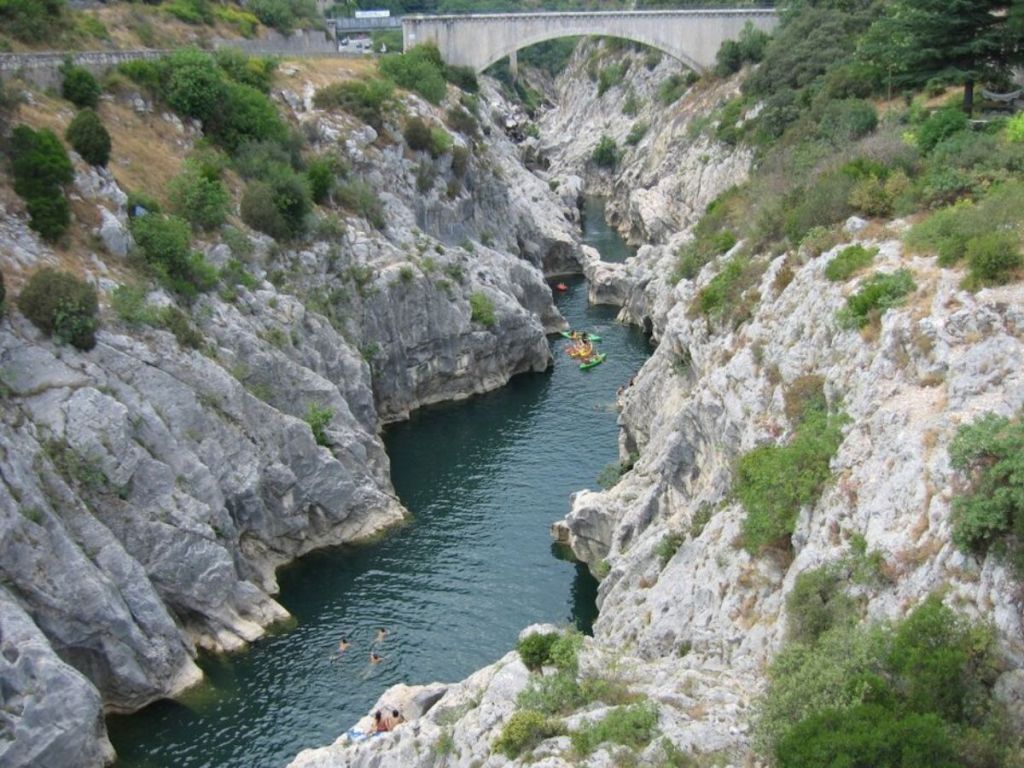 Aerial view of kayakers navigating the deep turquoise waters of the Hérault Gorges, flanked by dramatic white limestone cliffs and a stone arch bridge in the background