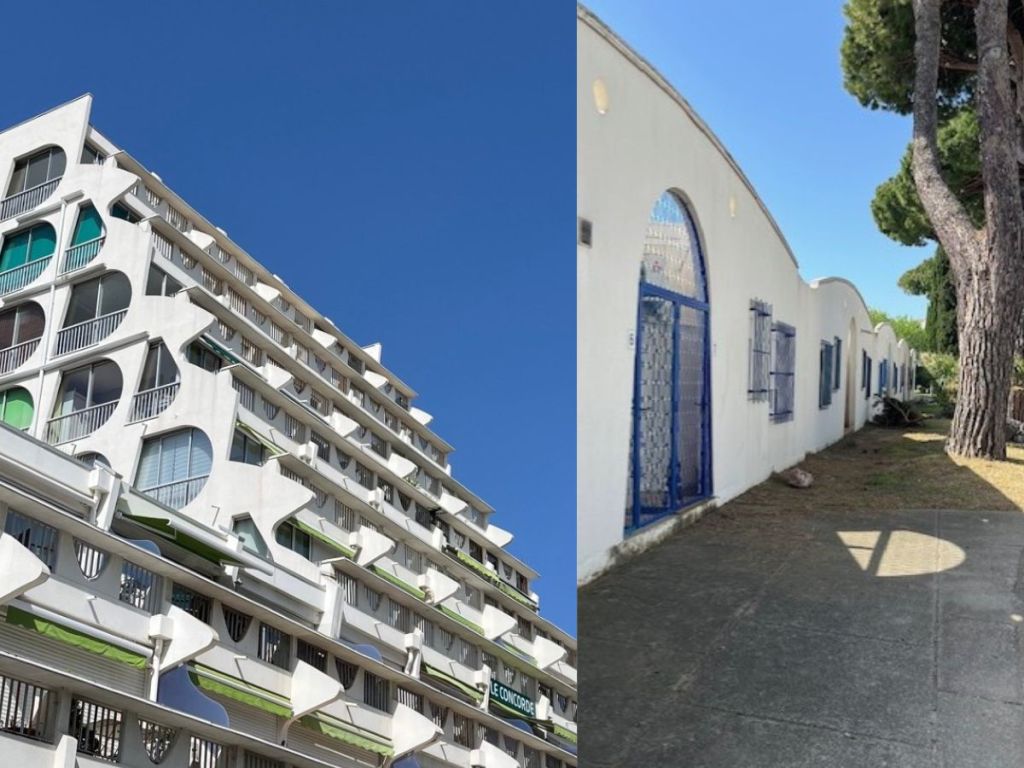Upward view of La Grande-Motte's iconic white brutalist apartment tower with rounded balconies against a deep blue sky, alongside a low white building with blue-trimmed arched doorways and umbrella pines.