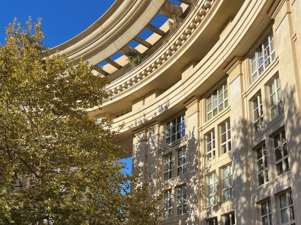 The curved neoclassical façade of a building in Montpellier's Antigone district, designed by architect Ricardo Bofill in the 1980s, with tall windows, decorative cornices, a rooftop pergola, and a plane tree in autumn foliage
