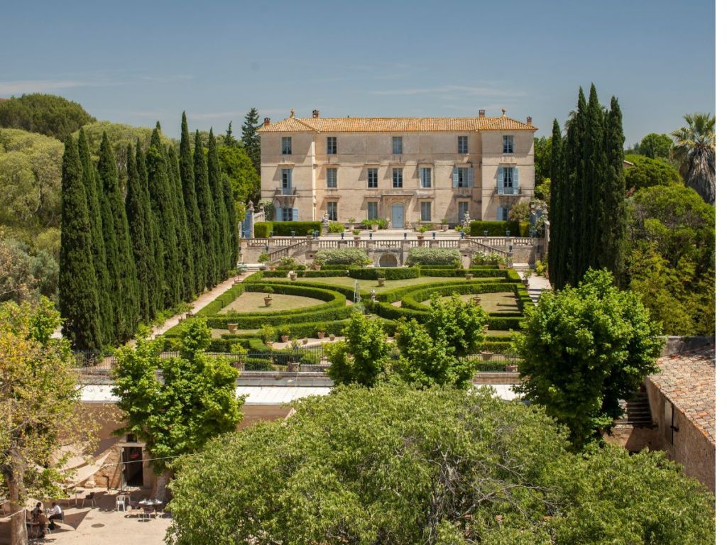 Aerial view of the Château de Flaugergues near Montpellier, an elegant 17th-century Italianate manor house surrounded by formal French gardens with geometric boxwood hedges, terracotta roof tiles, and a row of tall Italian cypresses under a bright blue sky
