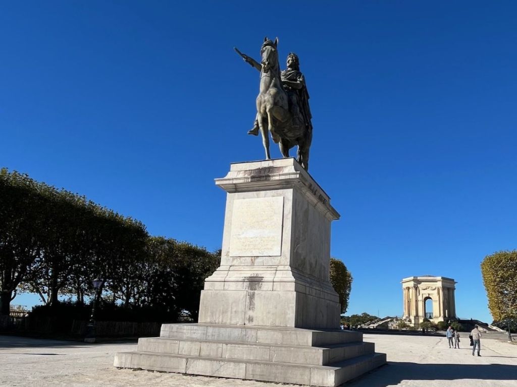 Bronze equestrian statue of Louis XIV on a marble pedestal at the Place Royale du Peyrou in Montpellier, with the 18th-century water tower and Saint-Clément aqueduct visible in the background under a cloudless blue sky