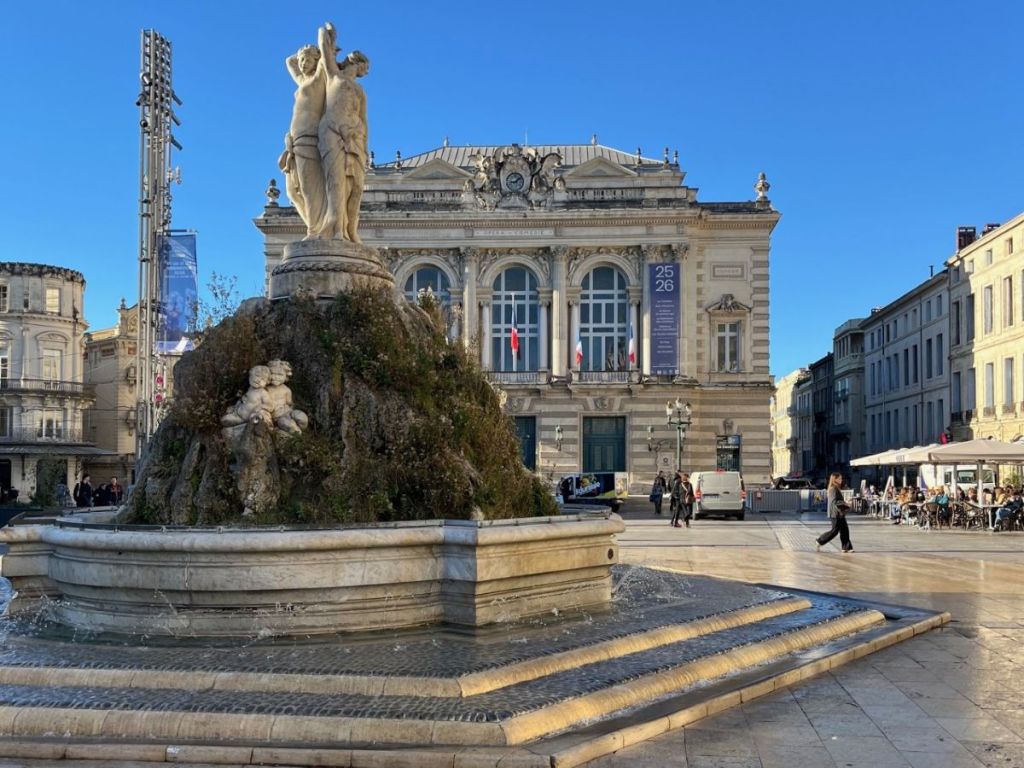 Place de la Comédie in Montpellier, a grand 19th-century square with the Opéra Comédie façade, the Three Graces fountain, and the city's iconic tramway curve on a sunny day