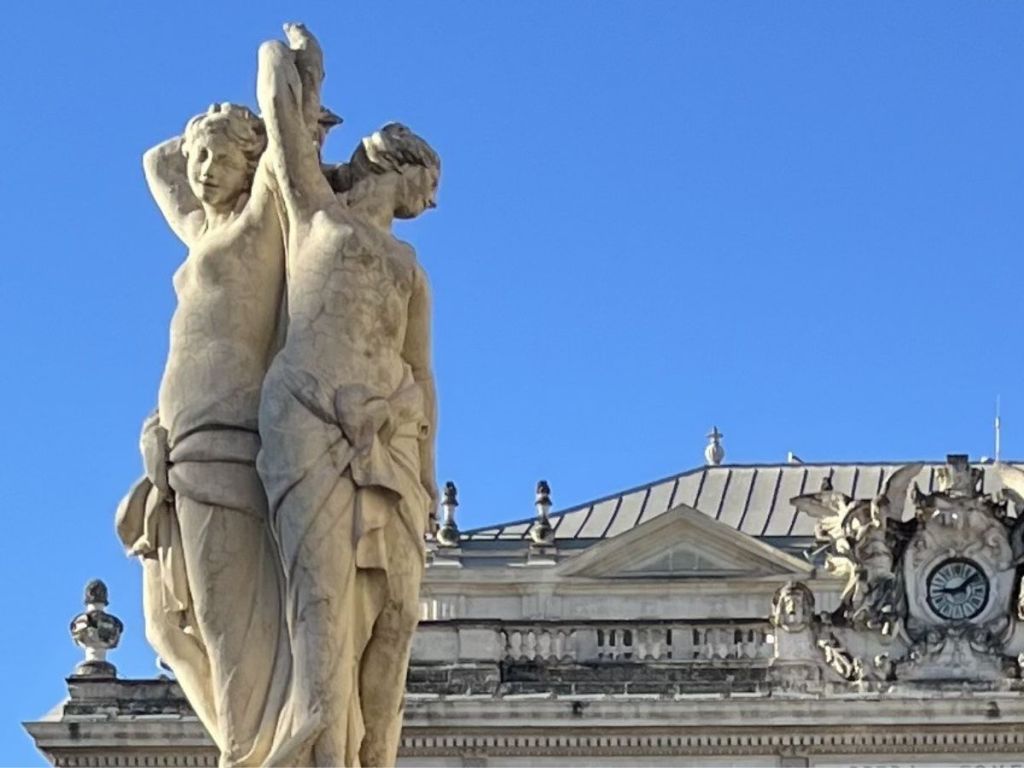 The Three Graces marble fountain at the center of Place de la Comédie in Montpellier, with the neoclassical Opera House façade rising behind it under a vivid blue sky