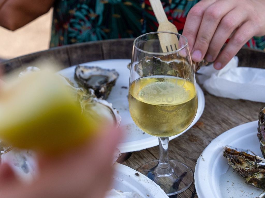 Close-up of a glass of white wine surrounded by freshly opened oysters on a wooden table, evoking a casual seaside tasting on the Thau Lagoon