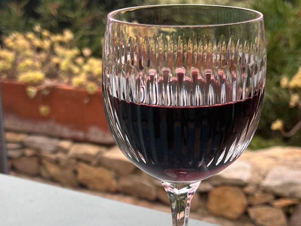 A crystal glass of deep red Pic Saint-Loup wine on a terrace, with a stone wall and Mediterranean garden in the background, Hérault.